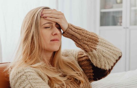 A close-up portrait of a distressed woman wearing a brown and dirty white knitted sweater, seated with her head resting in her hands showing signs of seasonal fatigue and headache.
