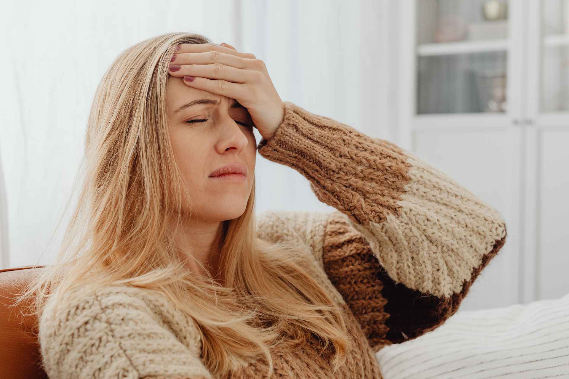 A close-up portrait of a distressed woman wearing a brown and dirty white knitted sweater, seated with her head resting in her hands showing signs of seasonal fatigue and headache.
