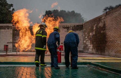 Three trainees dressed in full fire-resistant protective gear using a portable fire extinguisher to suppress controlled flames inside a live fire simulator setup.