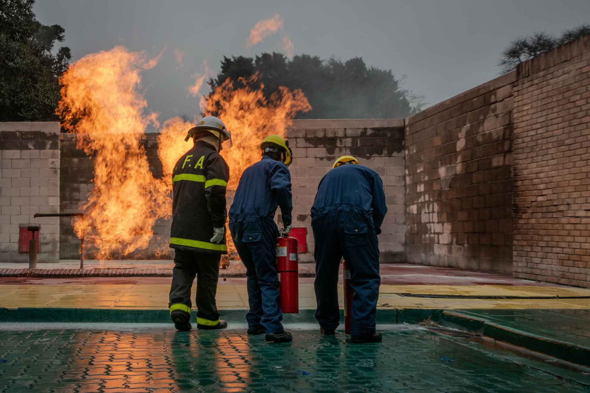 Three trainees dressed in full fire-resistant protective gear using a portable fire extinguisher to suppress controlled flames inside a live fire simulator setup.